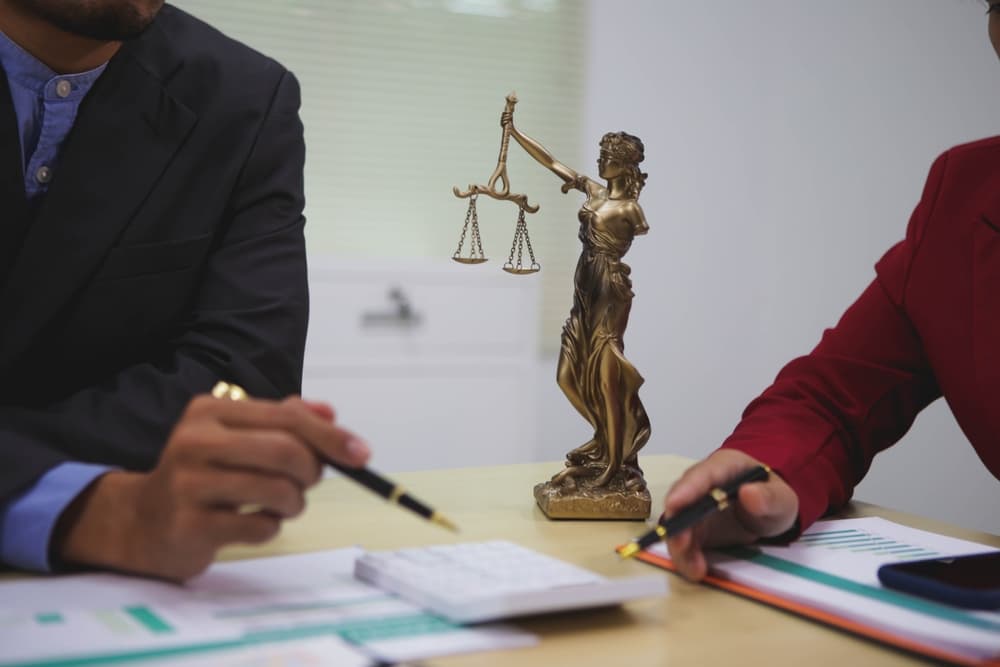 Two professionals discussing legal matters at a desk with Lady Justice statue, documents, and pens.