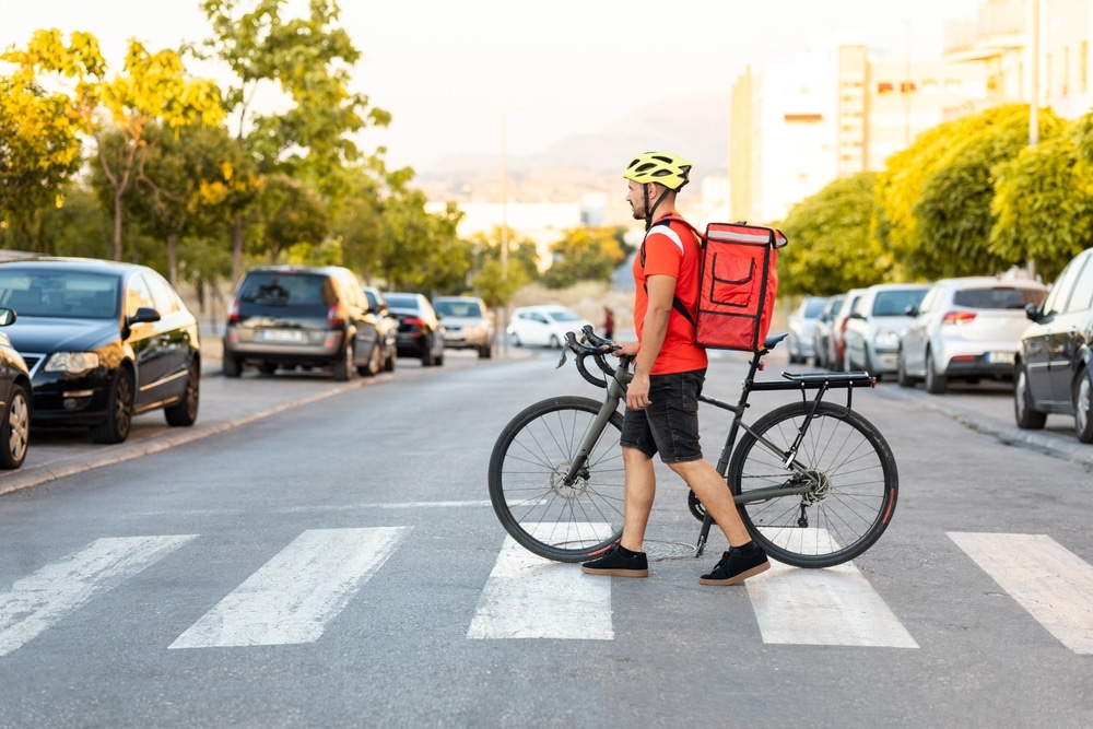 Delivery cyclist wearing a helmet and backpack crossing a city crosswalk near parked cars